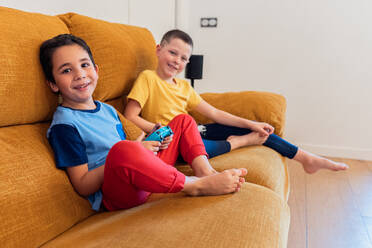Two young boys relaxing on an orange sofa while playing with game controllers in a cozy living room - ADSF54336