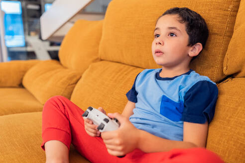 Junge, der sich mit Videospielen beschäftigt, auf einer senffarbenen Couch sitzend, mit einem Controller in der Hand - ADSF54339