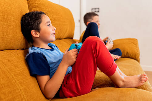 Zwei kleine Kinder spielen Videospiele auf einem gemütlichen orangefarbenen Sofa, wobei ein Kind einen blauen Controller hält - ADSF54341