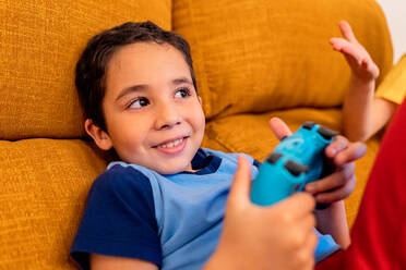 Lächelndes kleines Kind, das mit einem tragbaren Videospiel-Controller spielt, auf einer leuchtend gelben Couch sitzt und jemanden außerhalb des Bildes ansieht - ADSF54343