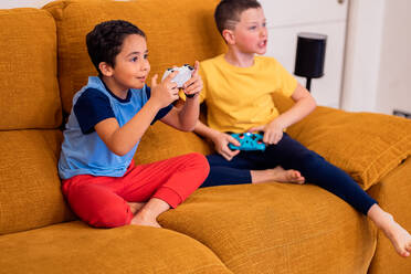 Two young boys happily engaged in playing video games, sitting on a luminous yellow sofa - ADSF54344