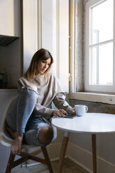 Frau in Freizeitkleidung, die am Fenster sitzt und nachdenkt, während sie ein Smartphone hält und eine Kaffeetasse auf dem Tisch steht - ADSF54347