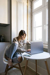 Eine junge Frau sitzt am Fenster und arbeitet mit einer Tasse Kaffee an ihrem Laptop in einem gut beleuchteten Raum - ADSF54349