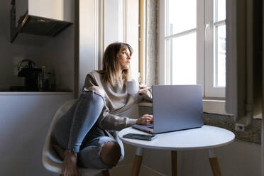 Eine Frau genießt einen ruhigen Moment mit einer Tasse Kaffee, während sie an ihrem Laptop in einer gemütlichen Umgebung zu Hause arbeitet, die durch das Fenster in natürliches Licht getaucht ist - ADSF54350