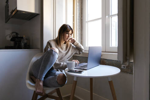 Eine junge Frau sitzt an einem kleinen runden Tisch am Fenster und arbeitet mit einer Tasse Kaffee nachdenklich an ihrem Laptop - ADSF54351