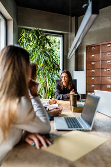 Zwei berufstätige Frauen unterhalten sich während einer Geschäftsbesprechung in einem modernen Büro, wobei eine Frau in die Kamera schaut. - ADSF54353