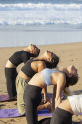 Die Teilnehmer machen Yoga-Rückbeugen am Strand während einer friedlichen Sitzung und verkörpern so Wellness und Harmonie mit der natürlichen Umgebung. - ADSF54360