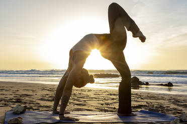 Ein Yoga-Praktizierender führt eine herausfordernde Pose an einem Sandstrand aus, während die Sonne untergeht und eine ruhige und inspirierende Umgebung schafft. - ADSF54363