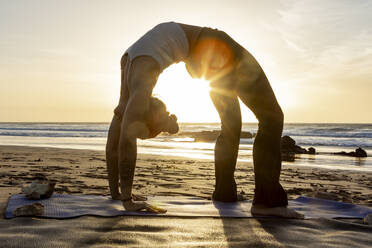 Üben einer fortgeschrittenen Yoga-Rückbeuge, der Rad-Pose, während eines ruhigen Sonnenuntergangs am Strand. - ADSF54364