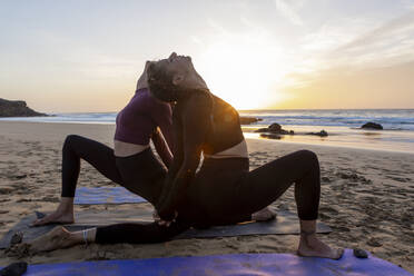 Two women practice yoga on a serene beach at sunset, each performing a distinct asana on their mats. - ADSF54365