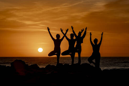 Eine Gruppe von Frauen nimmt an einer ruhigen Yogastunde am Strand teil, vor dem Hintergrund eines atemberaubenden Sonnenuntergangs, wobei jede eine andere Yogastellung einnimmt. - ADSF54367