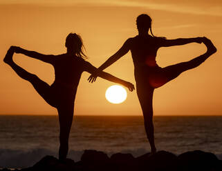 Zwei Silhouetten von Frauen, die eine gegenseitige Yogapose einnehmen, mit der untergehenden Sonne zwischen ihnen, die eine perfekte Mischung aus Gleichgewicht und Harmonie am Strand widerspiegelt. - ADSF54368