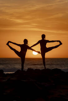 Zwei Silhouetten üben bei Sonnenuntergang am Strand Yogastellungen und fangen die Ruhe der Natur und die Harmonie der menschlichen Gestalt ein. - ADSF54369