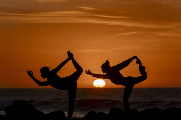 Zwei Frauen führen fortgeschrittene Yogastellungen auf Strandfelsen vor einer atemberaubenden Kulisse mit Sonnenuntergang aus. - ADSF54370