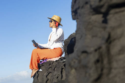 Seitenansicht eines digitalen Nomaden, der auf einem Felsen in einem sommerlichen Fischerdorf auf Fuerteventura sitzt, in seine Arbeit vertieft ist und telefoniert, während er das Küstenambiente genießt. - ADSF54373