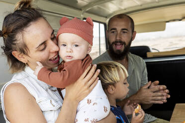 Eine herzerwärmende Szene einer Familie, die ihre Zeit in einem Retro-Van genießt, mit einer Mutter, die ihr Baby kuschelt, einem kleinen Jungen, der isst, und einem Vater, der mit Freude zusieht. - ADSF54379