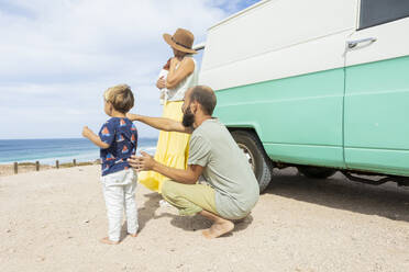 Eine junge Familie genießt einen ruhigen Moment am Meer neben ihrem Retro-Van und genießt die Freuden des einfachen Reisens und Zusammenseins. - ADSF54385