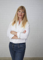 Beautiful natural blonde middle aged woman standing in front of white wall wearing white blouse and blue jeans with crossed arms - JBYF00267