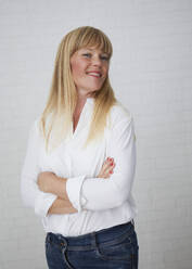 Beautiful natural blonde middle aged woman standing in front of white wall amiling and wearing white blouse and blue jeans with crossed arms - JBYF00268