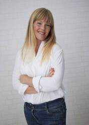 Beautiful natural blonde middle aged woman standing in front of white wall amiling and wearing white blouse and blue jeans with crossed arms - JBYF00269