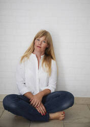 Beautiful natural blonde middle aged woman standing in front of white wall wearing white blouse and blue jeans sitting on floor - JBYF00275