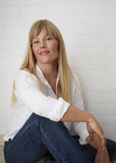 Beautiful natural blonde middle aged woman standing in front of white wall wearing white blouse and blue jeans sitting on floor - JBYF00276