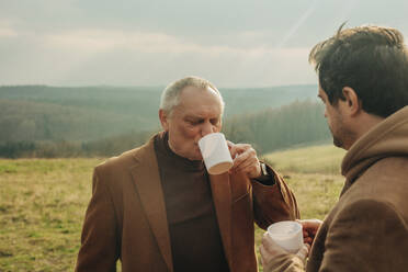 elderly father and middle-aged son drinkking tea in mugs in mountains and talking. Sudetes, Poland - VSNF01779