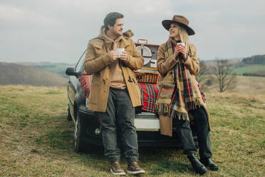 Stylish woman and man on picnic on car hood drinking tea in mugs. Moutains meadow, Sudetes, Poland - VSNF01785