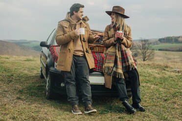 Stylish woman and man on picnic on car hood drinking tea in mugs. Moutains meadow, Sudetes, Poland - VSNF01786