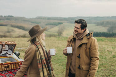 Stylish woman and man on picnic on car hood drinking tea in mugs. Moutains meadow, Sudetes, Poland - VSNF01787