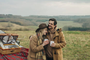 Stylish woman and man on picnic on car hood drinking tea in mugs. Moutains meadow, Sudetes, Poland - VSNF01789