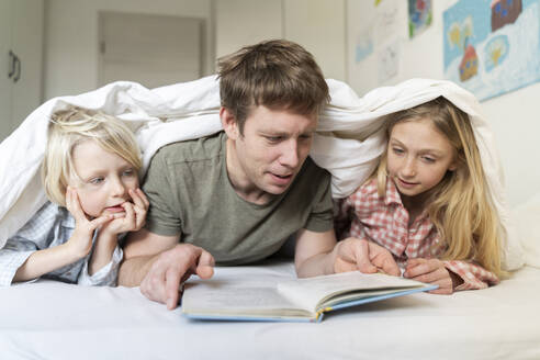 A father man with European appearance is reading book to his two children blond 6 years boy and 9 years old girl in pyjamas - NJAF00928
