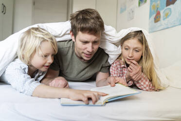 A father man with European appearance is reading book to his two children blond 6 years boy and 9 years old girl in pyjamas - NJAF00929