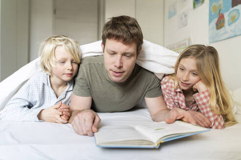 A father man with European appearance is reading book to his two children blond 6 years boy and 9 years old girl in pyjamas - NJAF00930