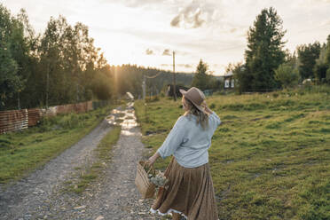 happy woman with a basket of herbs and flowers walks along a village road, a view from the back - VBUF00538
