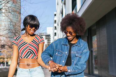 Two African American sisters walk hand in hand, their laughter and style shining brightly on a sunny city street - ADSF54408