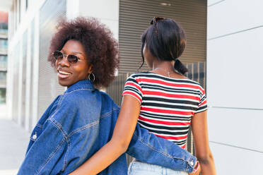 Looking over her shoulder, black woman with a bright smile walks arm in arm with her sister, showcasing urban fashion on a city street - ADSF54417