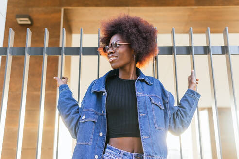 A confident woman in a denim ensemble exudes urban charm, her afro and sunglasses framing a joyful expression in the city - ADSF54421