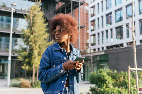 A fashion-forward woman pauses for a reflective moment with her smartphone amidst the bustling backdrop of modern urban architecture - ADSF54425