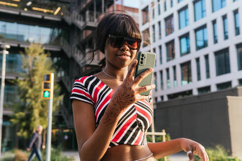 A young black woman with henna-adorned hand confidently takes a selfie while walking in an urban setting, showcasing her vibrant lifestyle - ADSF54426