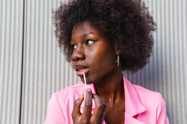 A fashionable black woman applies lip gloss with precision, her expression focused, highlighting a moment of personal style in an urban setting - ADSF54442