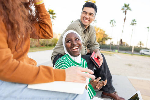 Diverse group of students with a laptop and smartphone enjoying a sunny day at a park, highlighting companionship and technology. - ADSF54457