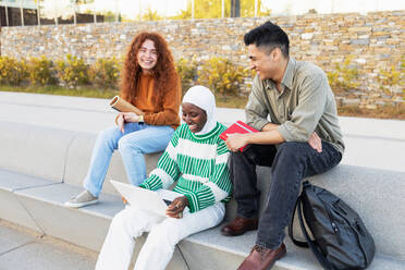 A smiling group of diverse students casually engaged outside with a laptop, books, and coffee, illustrating modern, inclusive education. - ADSF54458
