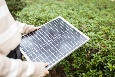 Close-up of person's hands holding a small solar panel above lush green bushes, representing sustainable energy. - ADSF54459