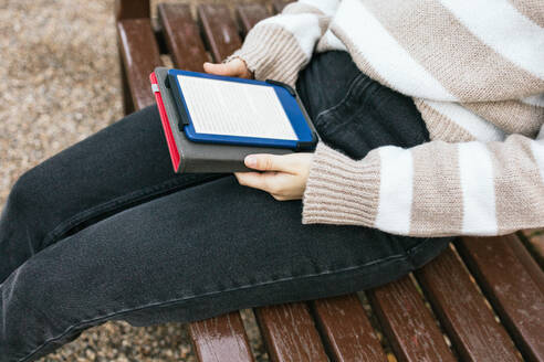 Close-up of an individual's lap while sitting on a bench with an e-book reader in hand, set against a casual outdoor backdrop. - ADSF54464