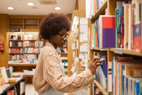 Side view of young black African American woman with glasses and a cozy sweater is thoughtfully selecting books from the shelves in a well-lit library - ADSF54465