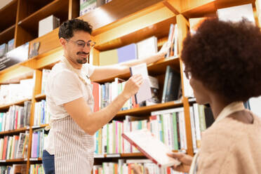 A young man hands a book to a African American woman amidst the aisles of a well-stocked library - ADSF54466