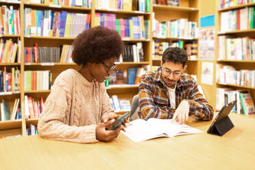 Two young students are engaged in a study session at a library, with books, a digital tablet, and smartphone - ADSF54471