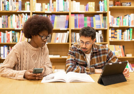 Two students engaged in studying at a library table with books and digital tablets, amidst rows of bookshelves - ADSF54472