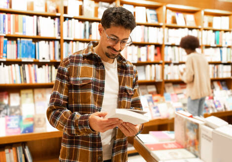 A young adult in a plaid shirt carefully selects a book from a shelf in a well-lit bookstore, surrounded by an extensive collection. - ADSF54474